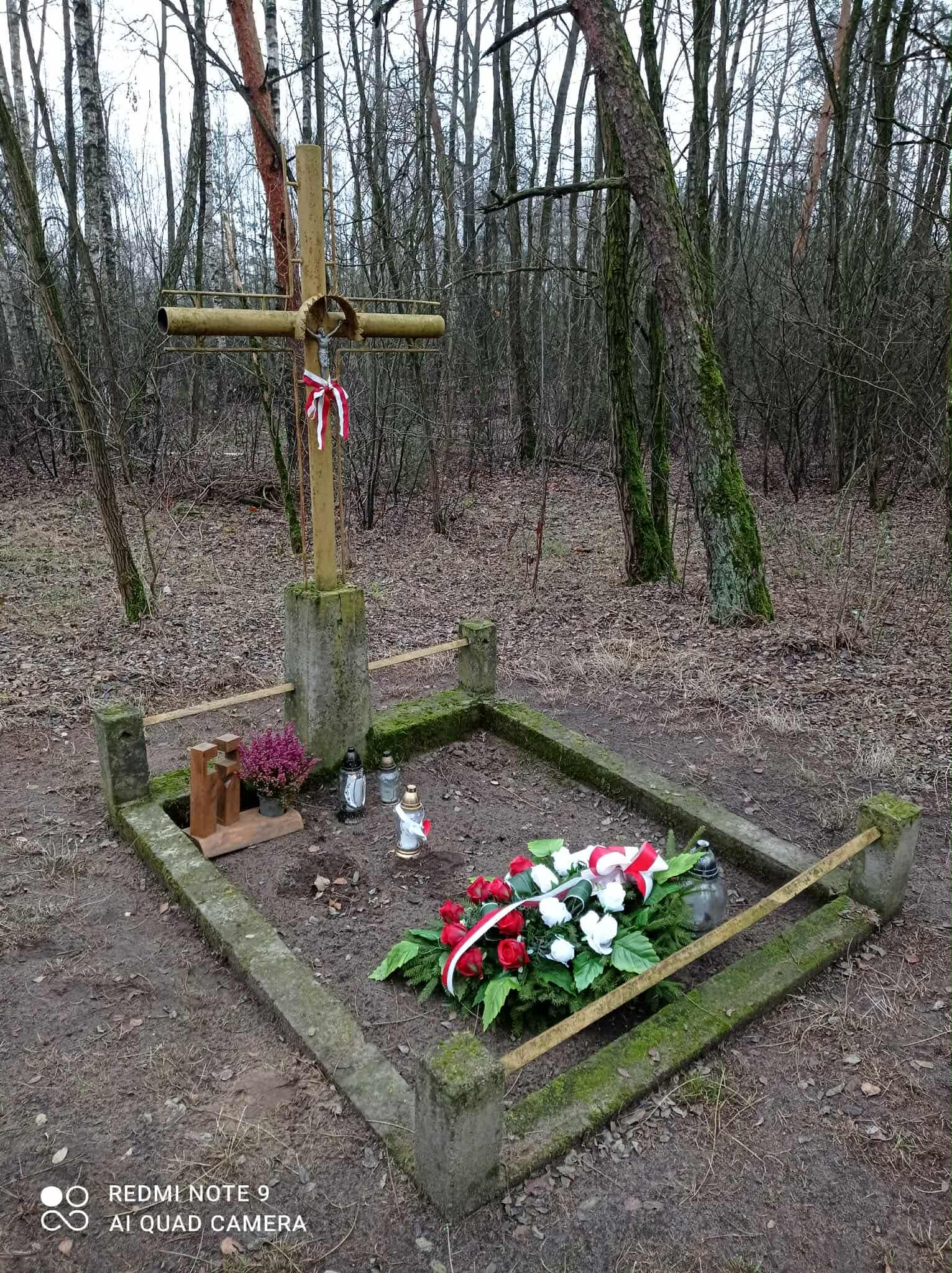Metalowy krzyż w lesie, na nim krucyfiks, bez żadnych inskrypcji. Betonowa podmurówka.