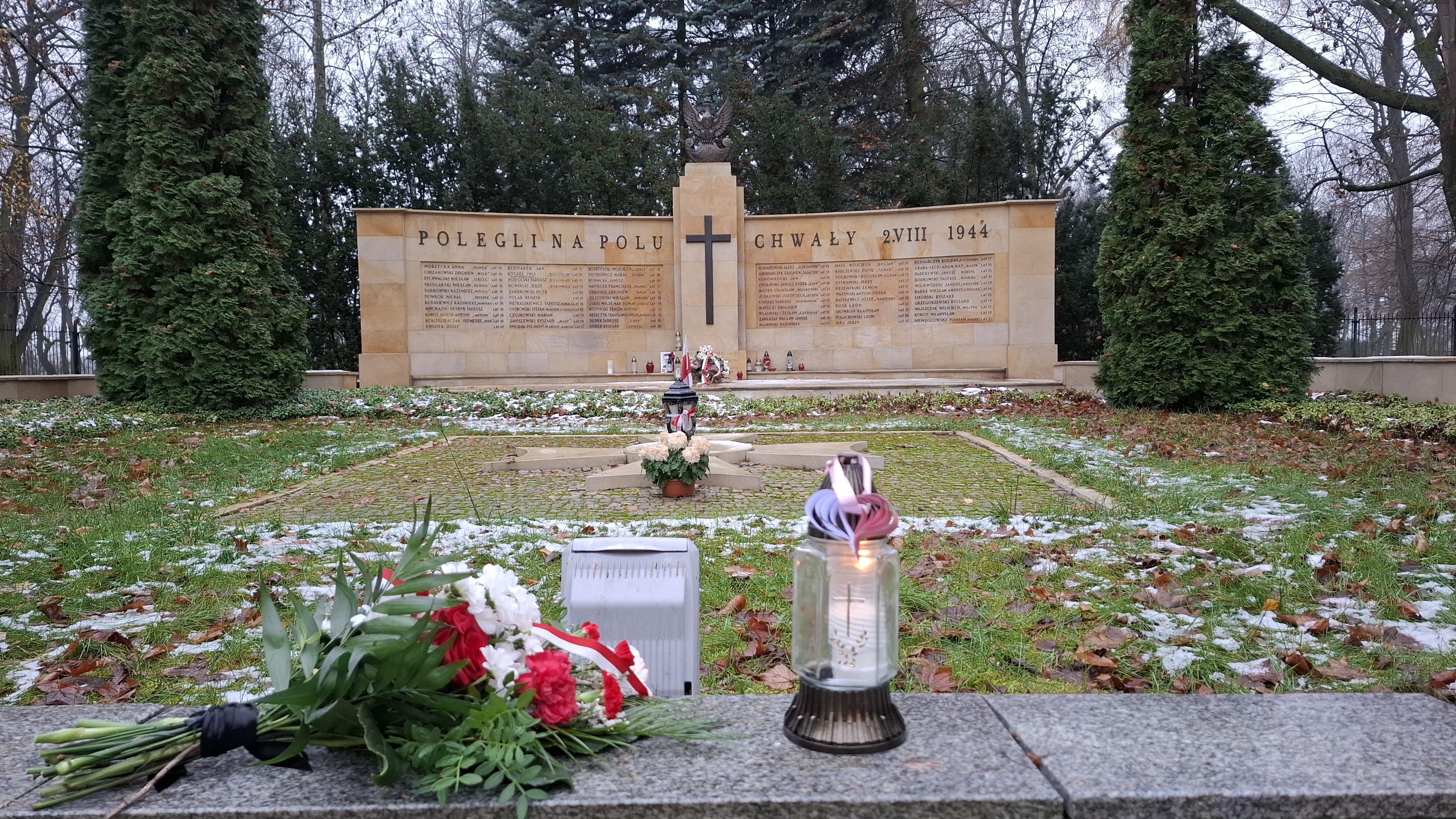 Znicz z zawieszonym papierowym biało-czerwonym sercem oraz bukiet kwiatów z biało-czerwoną wstążką