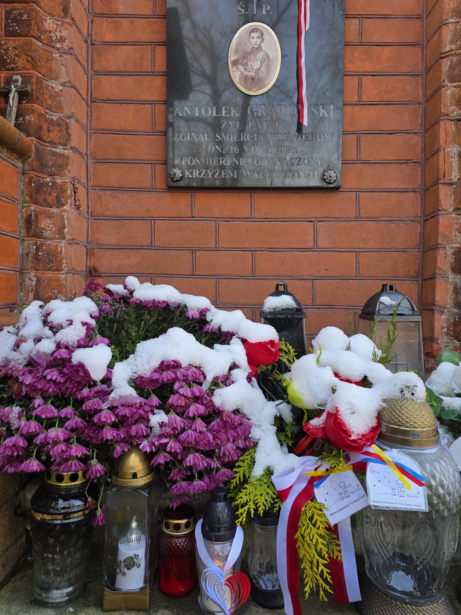 Znicz z zawieszką w kształcie serca na grobie, przed tablicą poświęconą bohaterowi